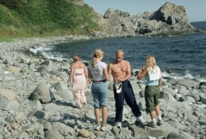 Wladimir Putin 2002 im Sommerurlaub mit seiner Familie: Zu sehen sind Putin von vorn und mit freiem Oberkörper sowie drei blinde Frauen unterschiedlichen Alters von hinten an einem steinigen Meeresstrand. / Foto © Imago/ITAR-TASS